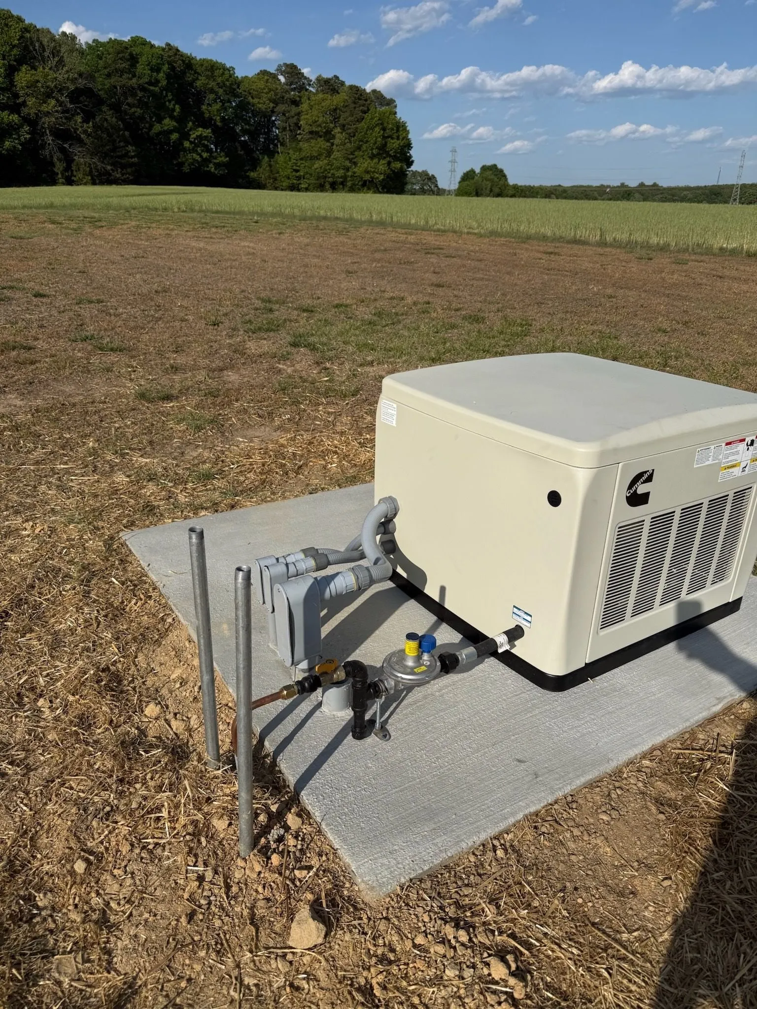 A new standby generator on a concrete pad at the edge of an open field, with its gas line and electrical conduit connected