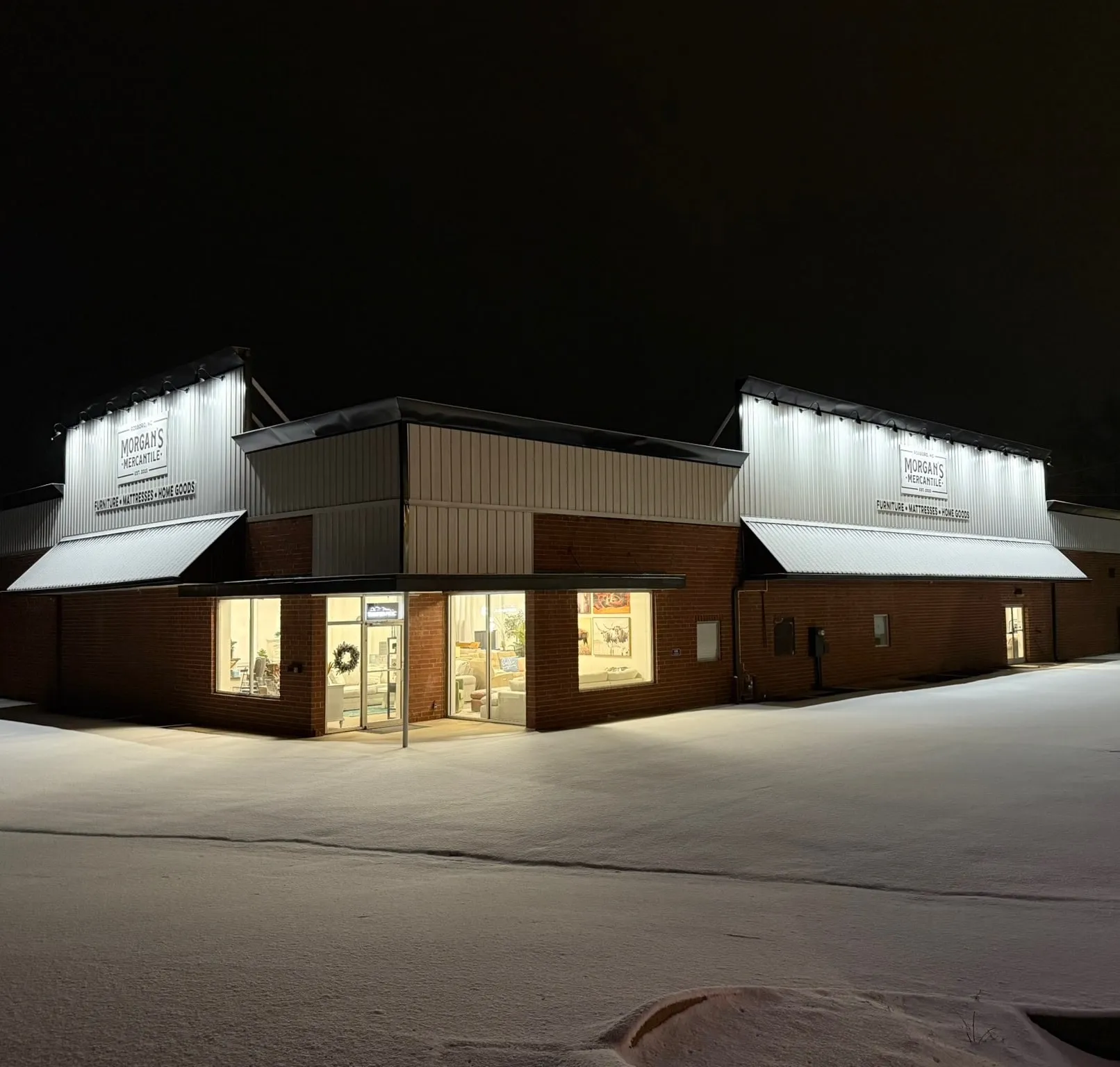 Morgan’s Mercantile at night after a snowfall, its two signs lit from above and the showroom windows glowing