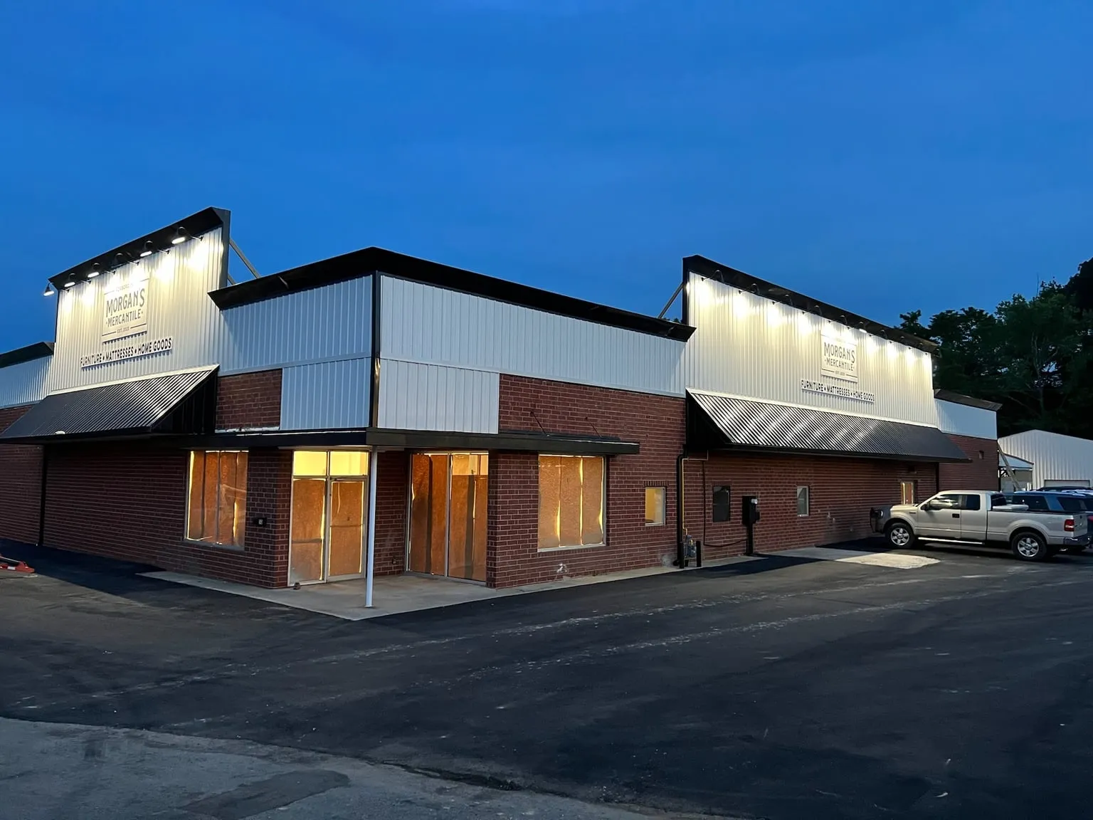 A brick and metal storefront at dusk, with a row of lights shining down on each of its two signs